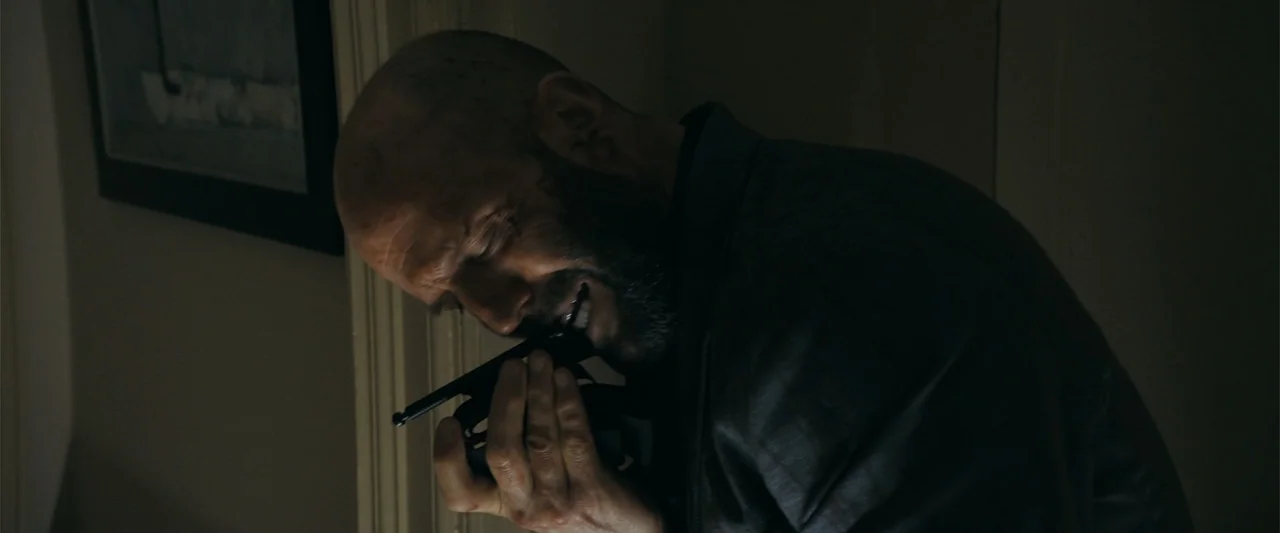The Beekeeper (2024) movie still: closeup — A dark indoor corner with minimal light cast upon a man's face. A man with a beard and shaved head holds…