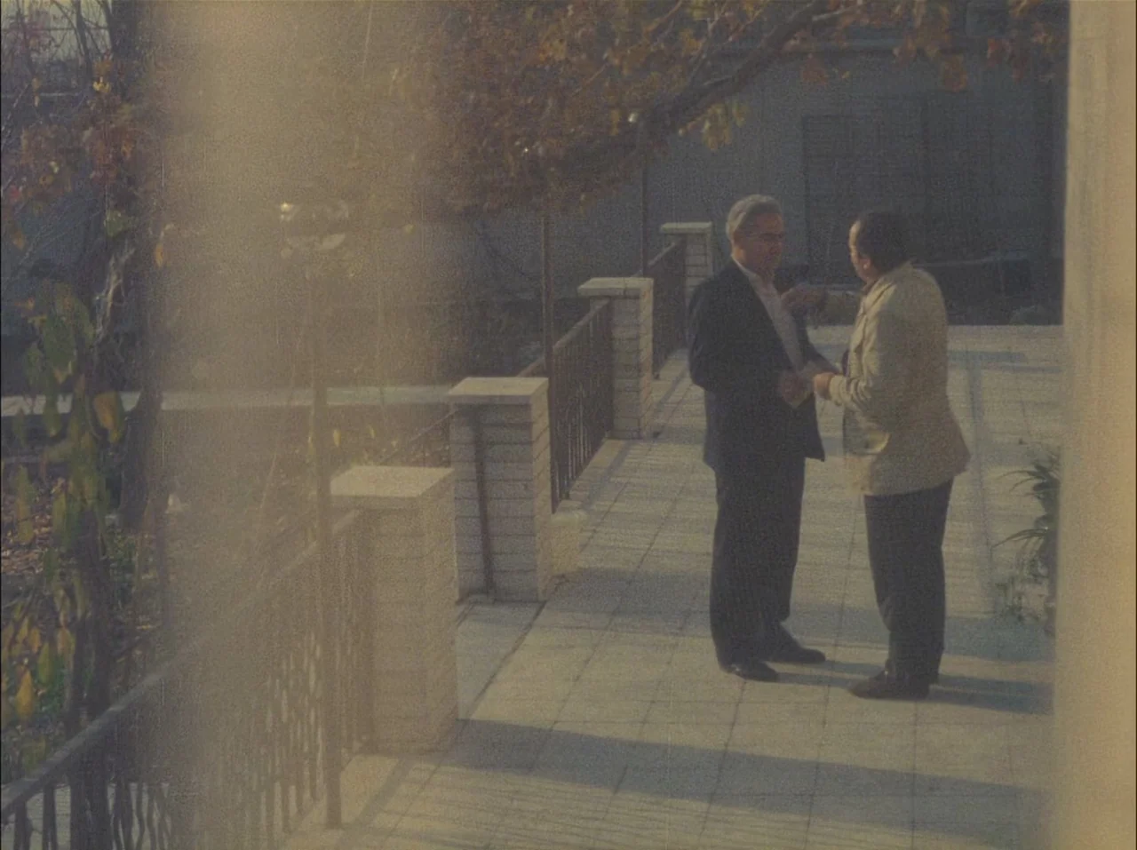 Close-Up (1990) movie still: fullbody — A stone terrace with metal railings overlooks a garden with autumn trees. Two middle-aged men stand on the…