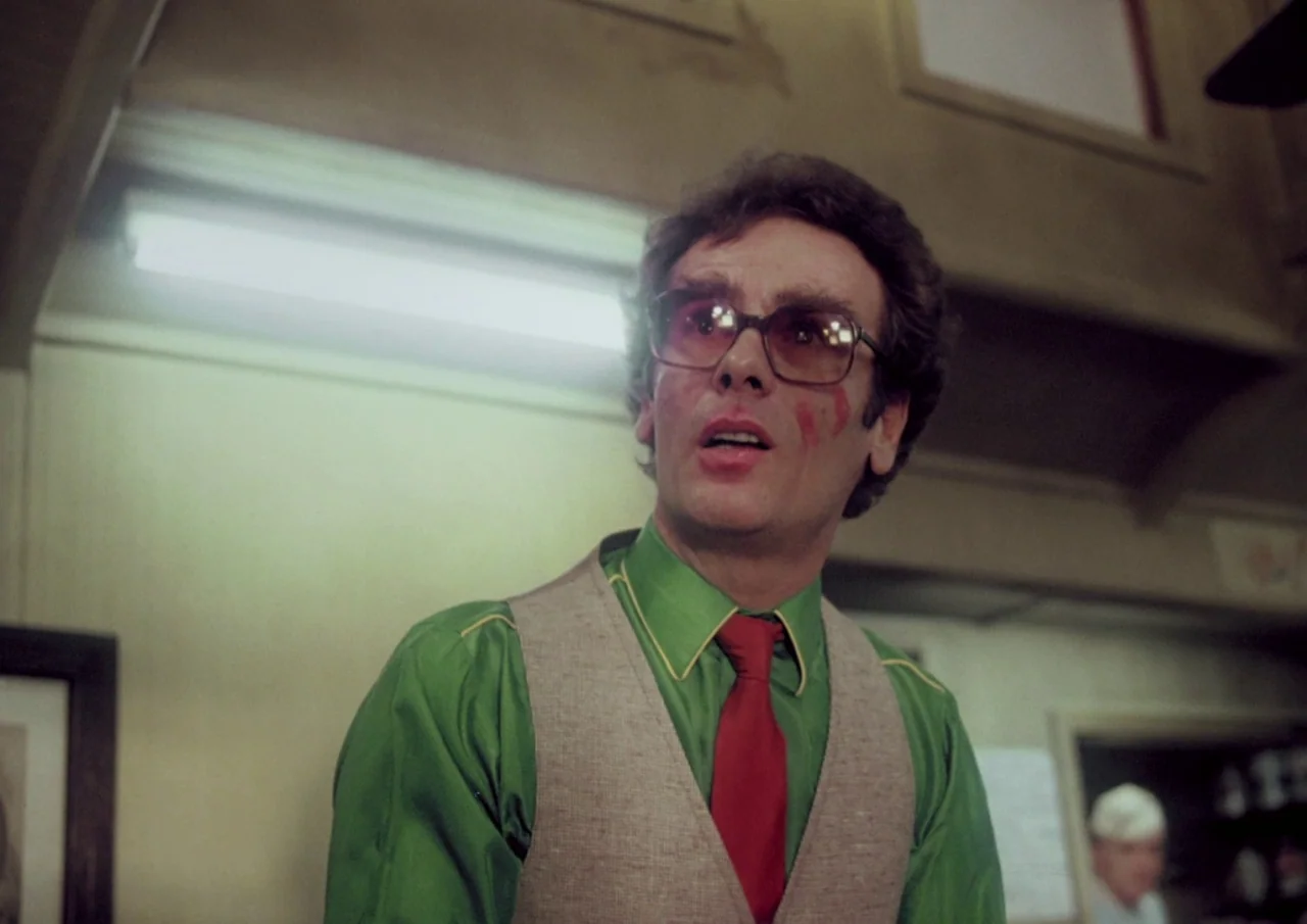 Human Highway (1982) movie still: medium — The man stands inside a brightly lit commercial diner setting with pale walls. He wears formal attire…