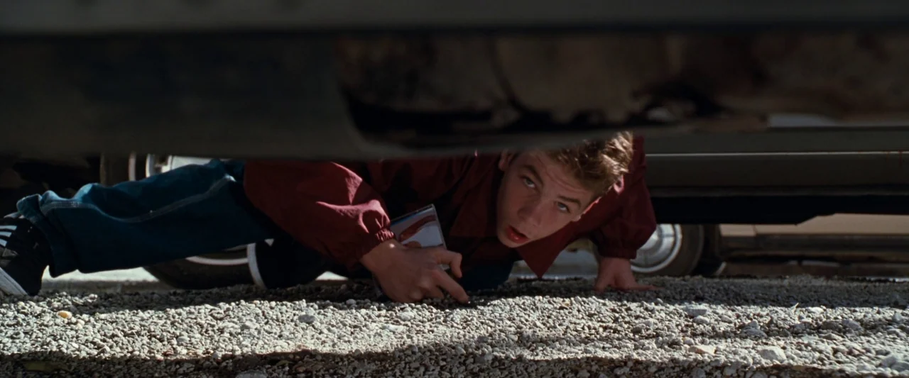 Empire Records (1995) movie still: medium — A teenage boy crawls underneath a car parked on a bed of loose gravel. He reaches toward the ground…
