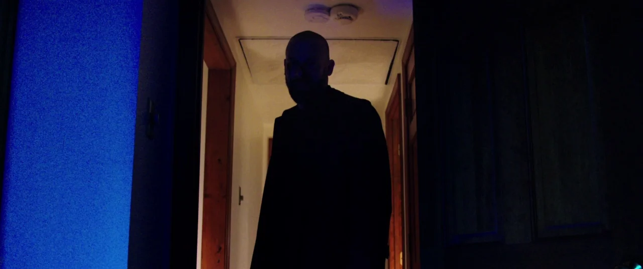 The Mind’s Eye (2015) movie still: medium — A dark residential hallway with wooden door frames leads into a dimly lit room. A man with a shaved head…