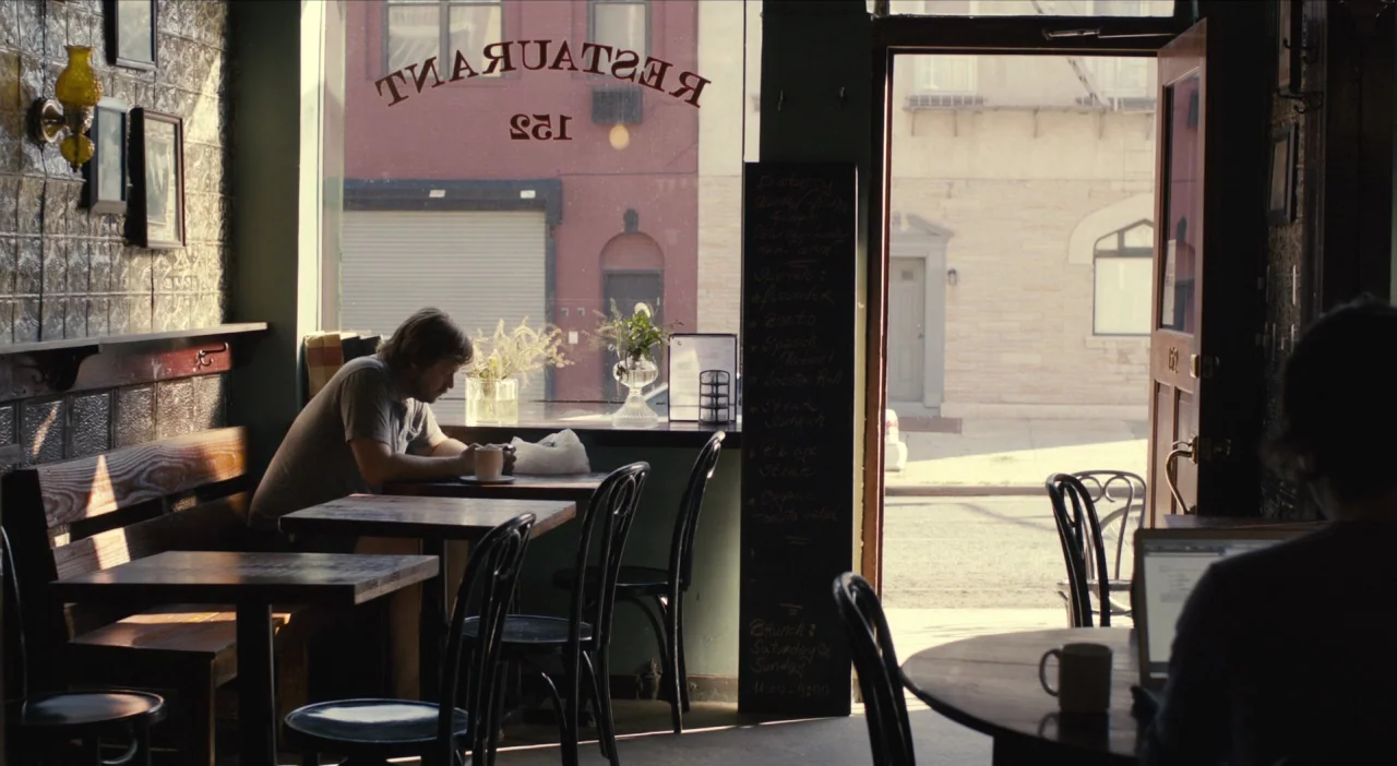 The Comedy (2012) movie still: medium — An interior shot of a small cafe with wooden tables and a chalkboard menu. A man in a t-shirt sits by the…