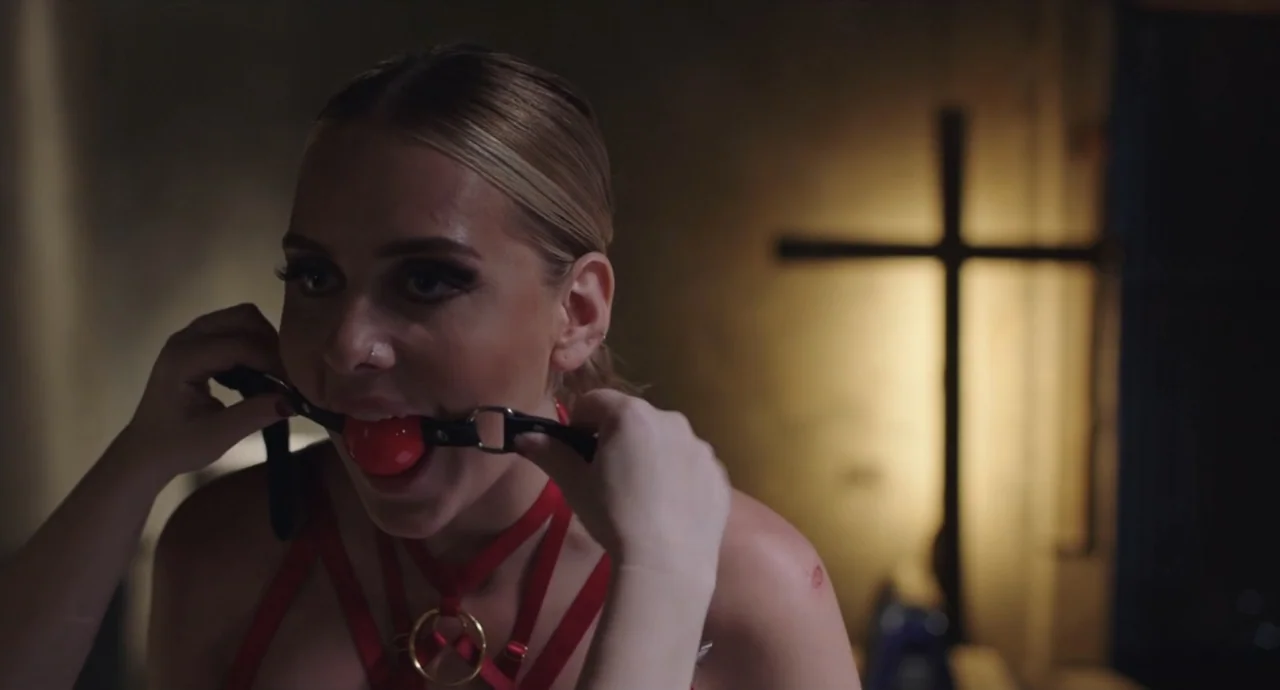 Pleasure (2021) movie still: closeup — A dimly lit indoor space featuring a blurred wooden cross standing in the background. A woman in a red…