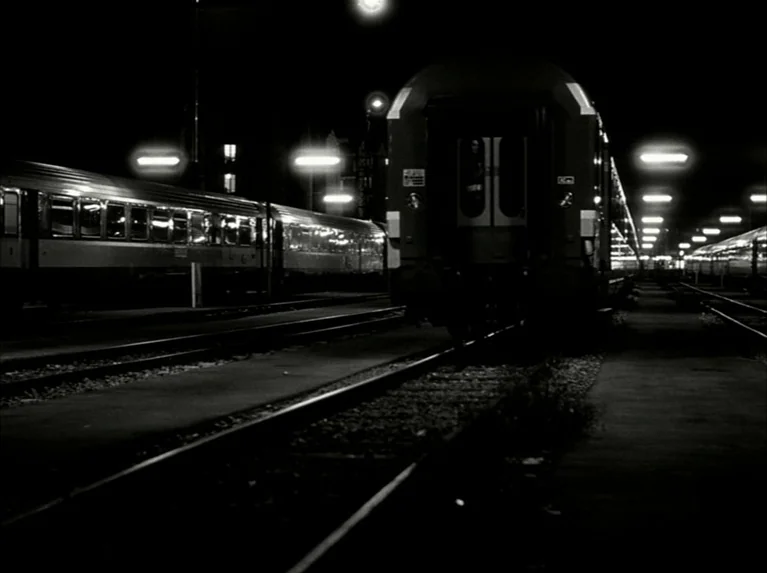 Eloge de L’amour (2001) movie still: wide — A night view of multiple passenger train cars parked on parallel sets of tracks. Rows of streetlights…