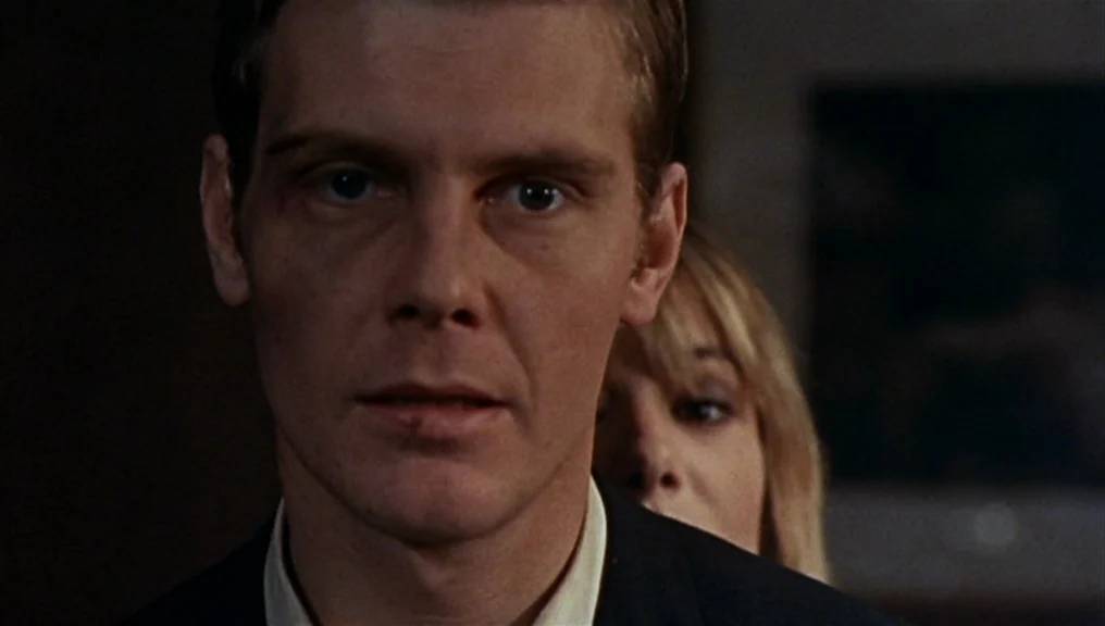 Performance (1970) movie still: closeup — A man stands in the foreground with a woman positioned behind his right shoulder. The background is…