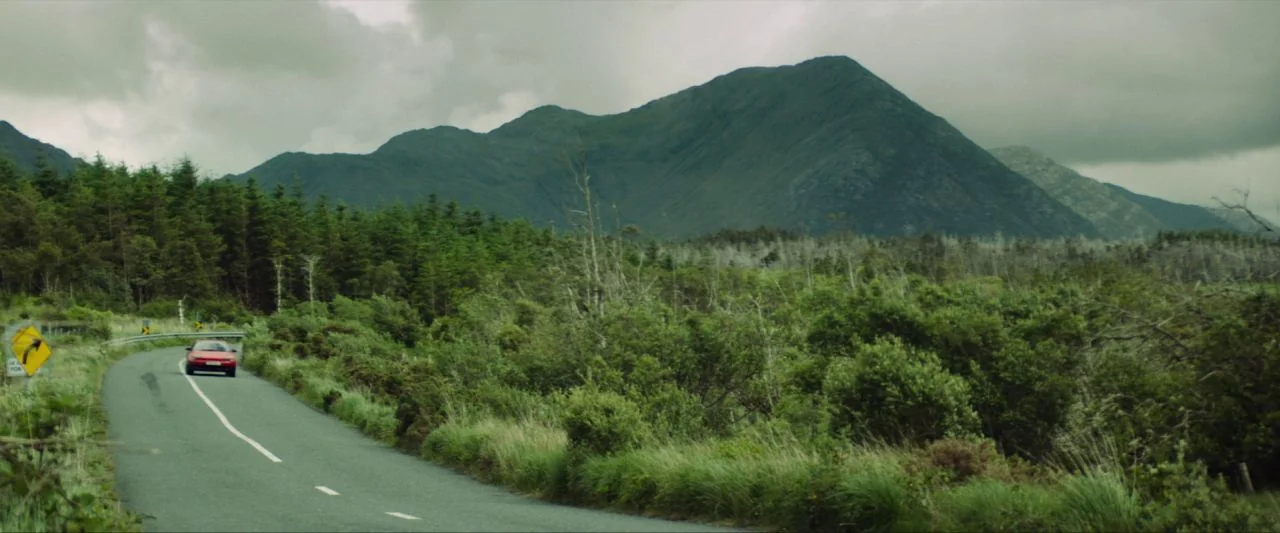 Calm With Horses (2019) movie still: wide — A rural asphalt road winds through a lush green valley beneath large mountains. A small red car travels…