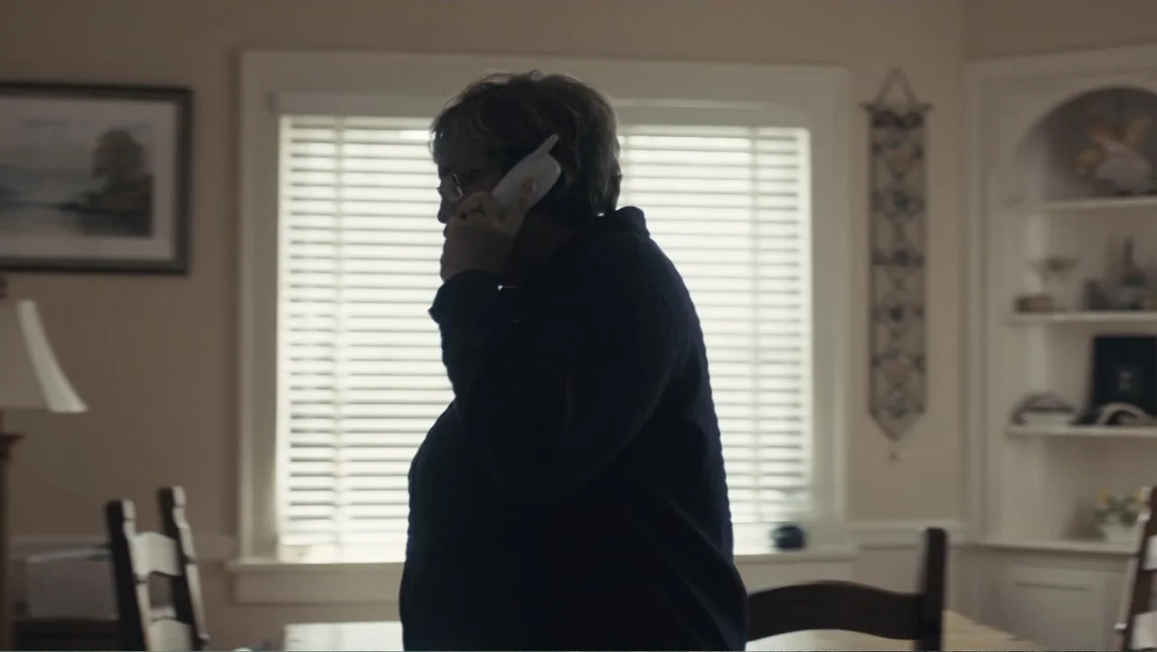 Paterno (2018) movie still: medium — A dining room features light-colored walls, a window with blinds, and built-in shelving. A woman in a dark…