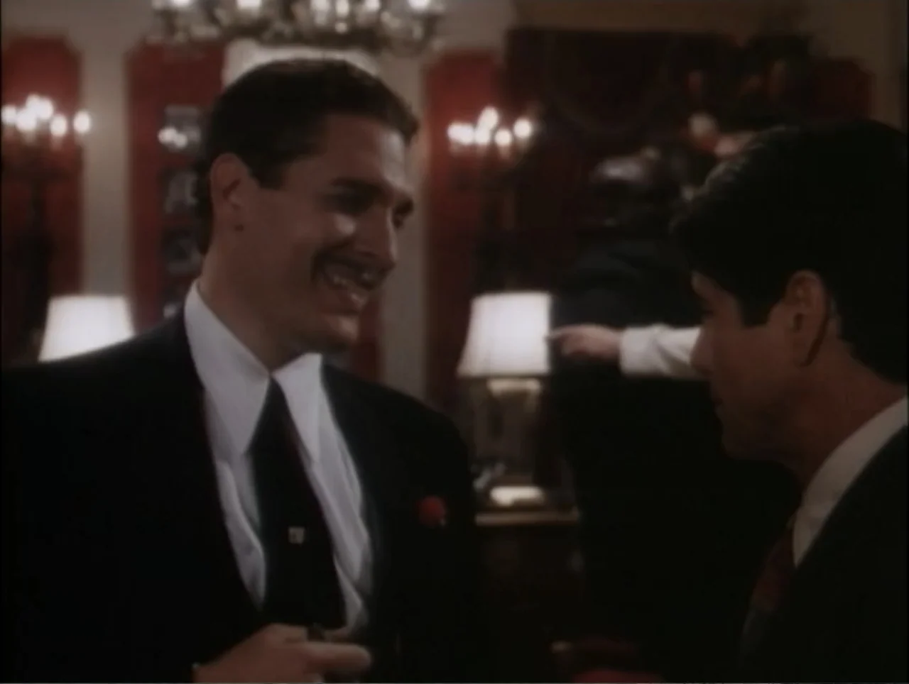 Cast a Deadly Spell (1991) movie still: medium — A dimly lit indoor event space features red wall panels and decorative ceiling lights. Two men in…