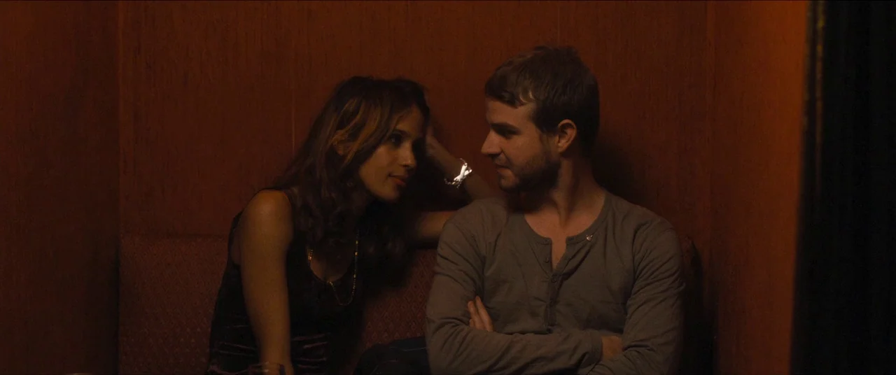 Simon Killer movie still: medium — A dimly lit wooden booth inside a bar or restaurant with warm tones. A man and woman sit opposite each other in a…