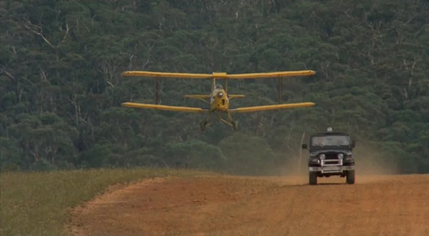 The Coca-Cola Kid (1985) movie still: wide — A yellow biplane flies directly over a dirt road in front of a dark jeep. A dense forest of green…