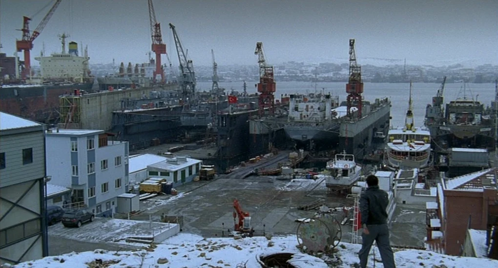 Uzak (2002) movie still: wide — A snow-dusted industrial shipyard features multiple cranes and dry-docked ships along the water. A man stands on a…