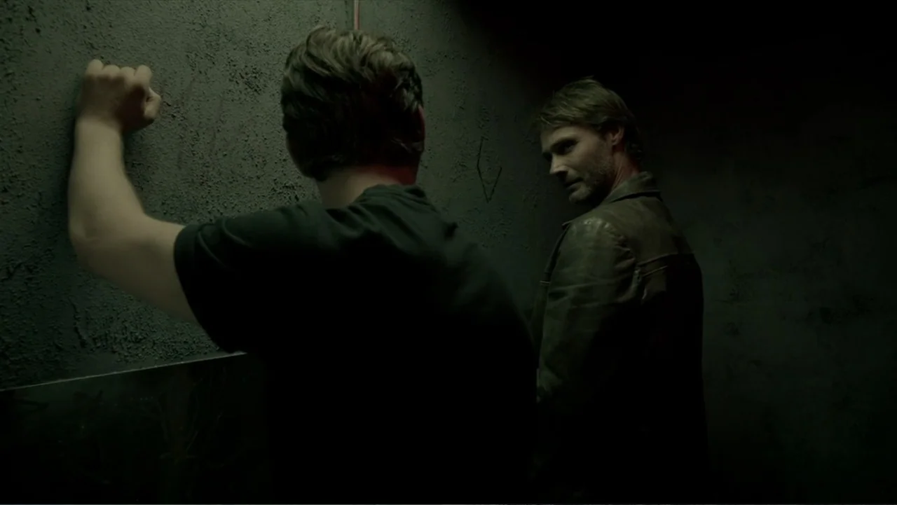 Copenhagen (2002) movie still: medium — A dark, cramped concrete room with textured walls and dim ambient illumination. One man leans against the…
