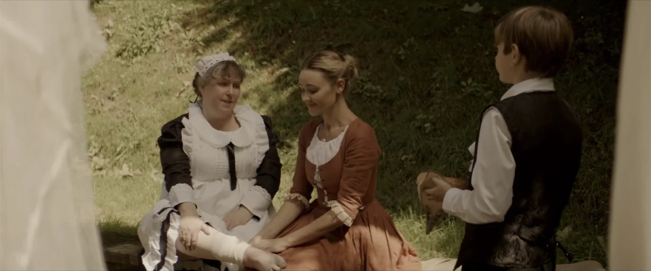 Cinderella's Curse (2024) movie still: medium — A grassy slope provides a setting for three people gathered near a wooden structure. A woman in a…