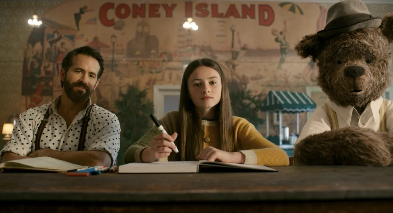 IF (2024) movie still: medium — A wooden table is positioned in front of a painted Coney Island mural backdrop. A man, a young girl with a marker,…