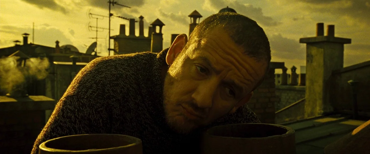 Micmacs (2009) movie still: closeup — A rooftop at sunset features several masonry chimney stacks with antennas in the background. A man leans…