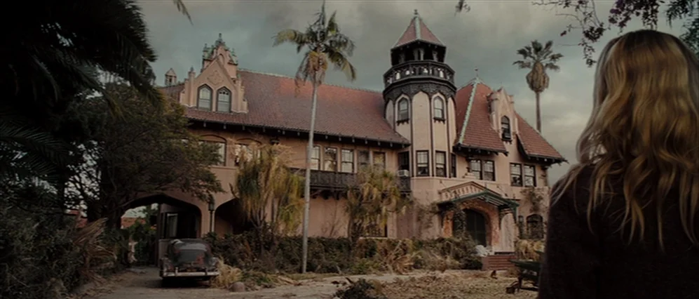 Drag Me to Hell (2009) movie still: wide — An ornate two-story stucco mansion with a corner turret sits beneath a cloudy sky. A woman stands in the…