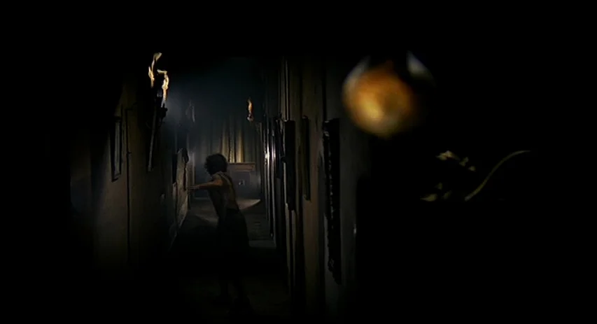 Malpertuis (1971) movie still: wide — A long, dark corridor is illuminated by wall-mounted torches casting long, dramatic shadows. A person in…