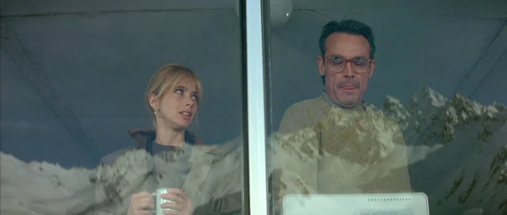 The Big Blue (1988) movie still: medium — A man and a woman stand behind a window reflecting snow-capped mountain peaks. The woman holds a mug while…