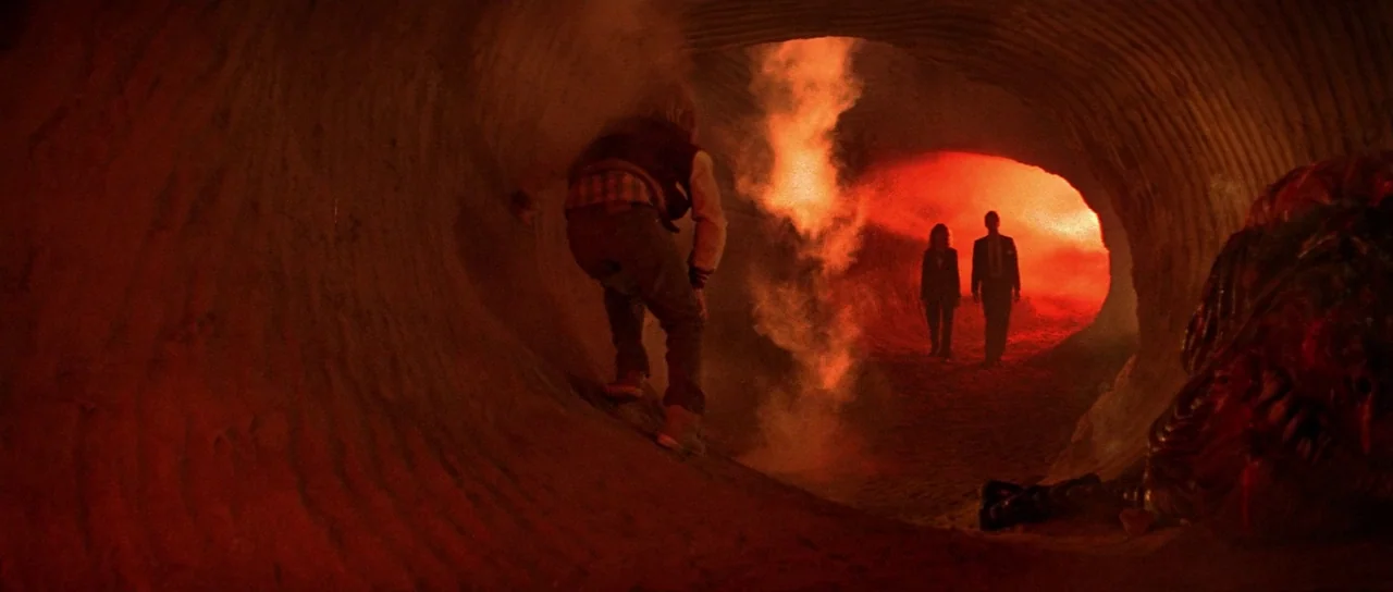 Invaders From Mars (1986) movie still: wide — A long corrugated metal tunnel is filled with thick orange smoke and red illumination. Three people…