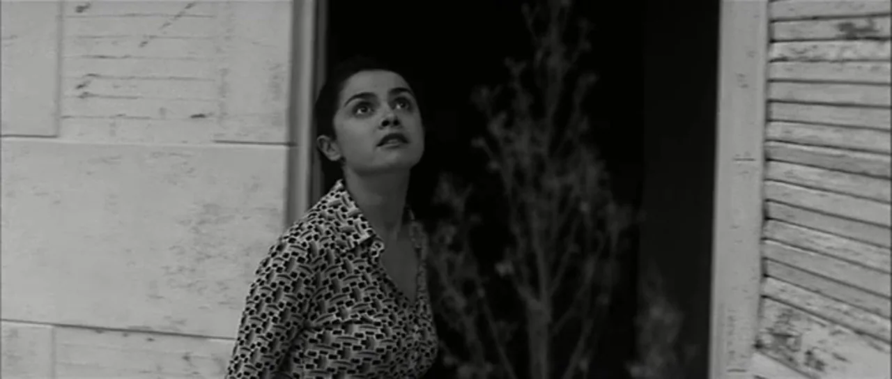 13 Tzameti (2006) movie still: medium — A stone doorway exterior features white textured wall panels and a dark recessed opening. A woman wearing a…