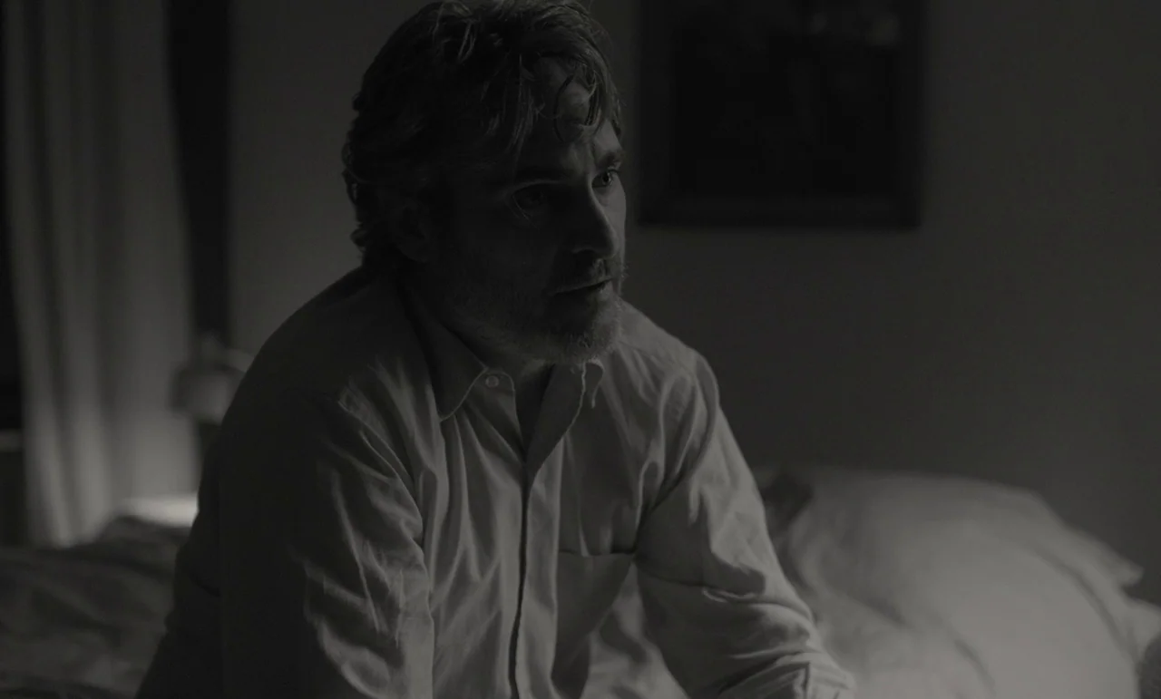C’mon C’mon (2021) movie still: medium — A dimly lit room with bedding visible in the foreground and a plain wall background. A middle-aged man with…