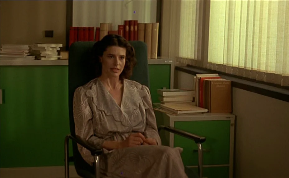 The Woman Next Door (1981) movie still: medium — A workspace features sage green cabinets, stacks of books, and closed venetian blinds. A woman sits…
