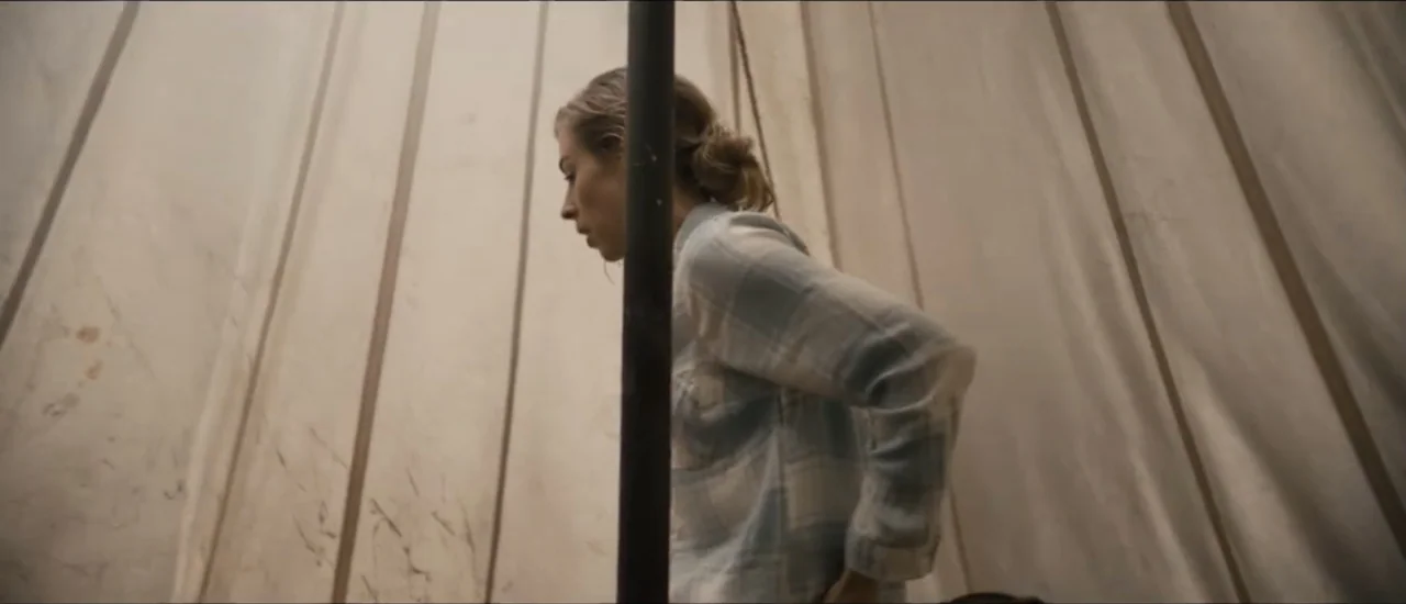 Fear Below (2025) movie still: medium — A confined space inside a canvas tent with vertical pole structures. A young woman in a plaid shirt stands…