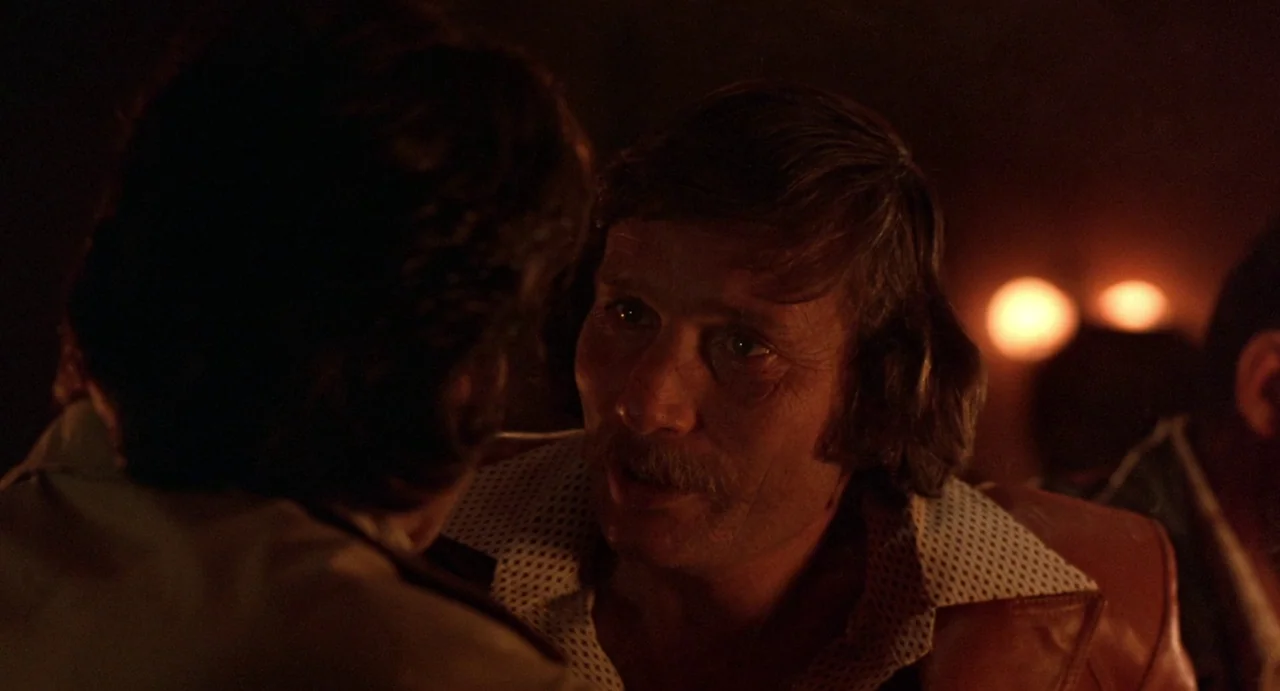 Straight Time (1978) movie still: closeup — A dimly lit room with warm light sources reflecting off wall textures. A mustachioed man faces another…