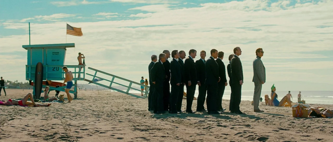 Pawn Sacrifice (2014) movie still: wide — A group of men in dark business suits stands on a sunny sandy beach. Behind them a wooden lifeguard tower…
