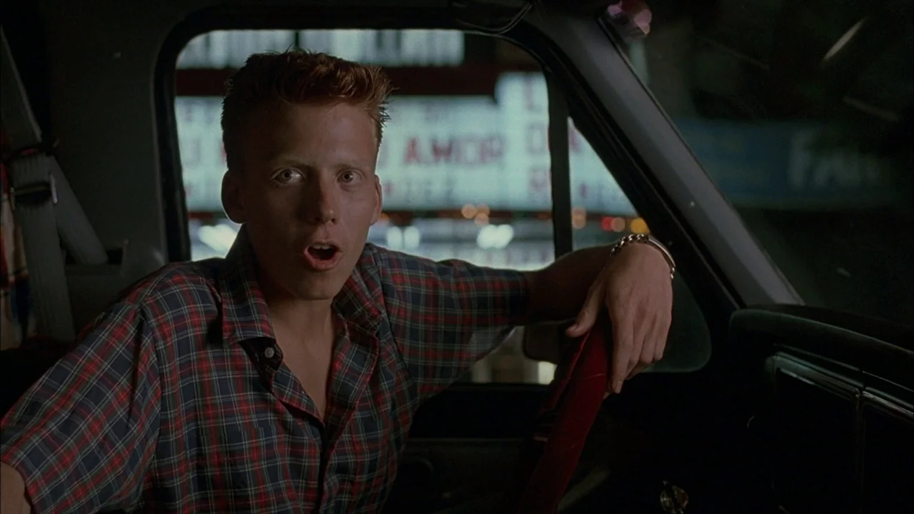 Whore (1991) movie still: medium — A man sits behind the wheel of a car at night with city lights visible outside. The driver looks forward with an…