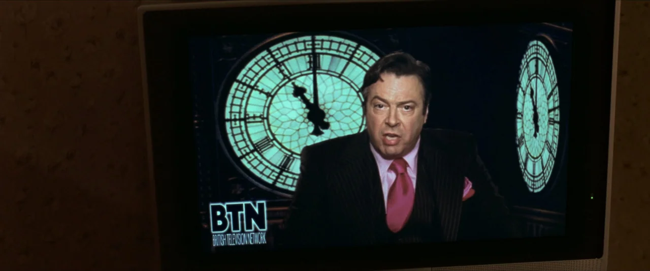 V for Vendetta (2006) movie still: medium — A televised news anchor is framed by large glowing clock faces in a broadcast graphic. The man wears a…