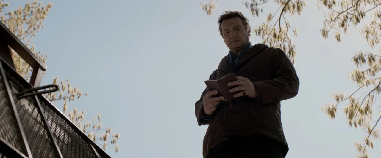 A Walk Among the Tombstones (2014) movie still: medium — An outdoor park area with bare tree branches silhouetted against a pale sky. A mature man…