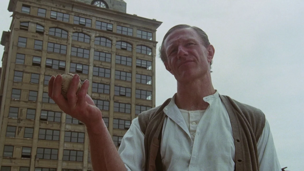 Insignificance (1985) movie still: medium — An urban setting features a tall, weathered brick building against a pale overcast sky. A man stands in…