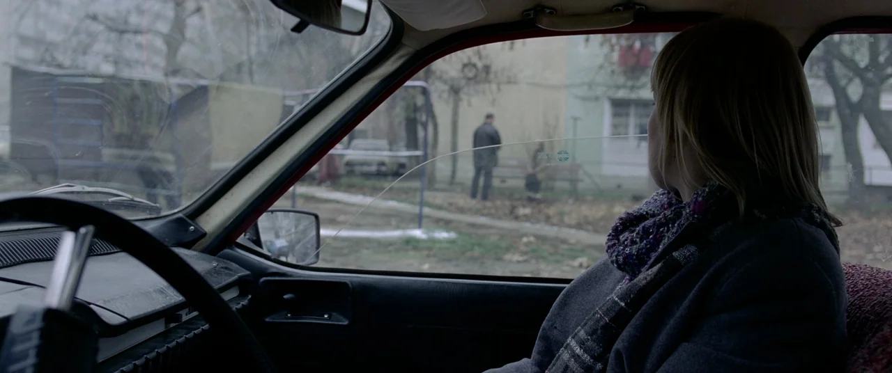 4 months, 3 Weeks and 2 Days (2007) movie still: medium — A woman sits inside a car looking out at a residential exterior. A lone man stands on a…