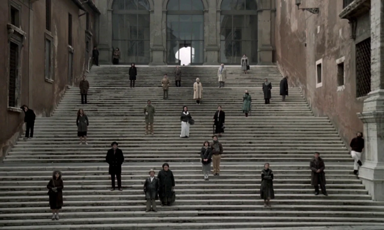 Nostalghia (1983) movie still: wide — A wide stone staircase sits between two tall brown stone buildings. A group of people are scattered at various…