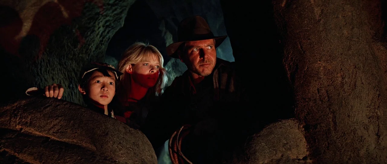 Indiana Jones and the Temple of Doom (1984) movie still: medium — Three people hide behind large dark rocks inside a dim, textured natural cave. The…
