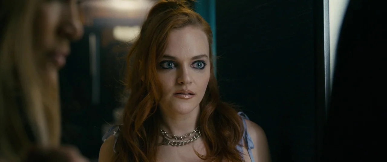 Hustlers (2019) movie still: medium — A dark, dimly lit hallway serves as the backdrop for the interaction. A red-headed young woman stands near a…