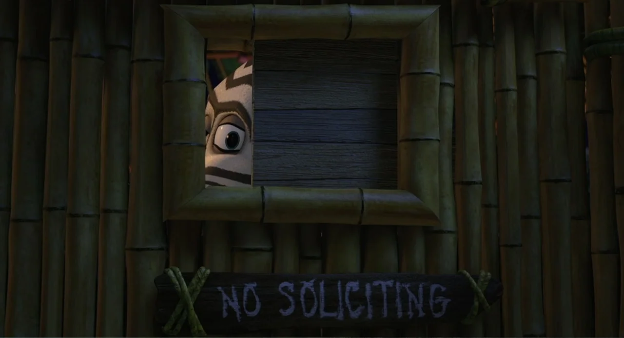 Madagascar (2005) movie still: closeup — A vertical bamboo wall features a small rectangular window and a rustic wooden sign. A striped creature…
