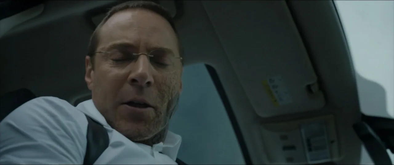 Kraven the Hunter (2024) movie still: closeup — A man sits inside a vehicle with daylight streaming through the window. The man has his eyes closed…
