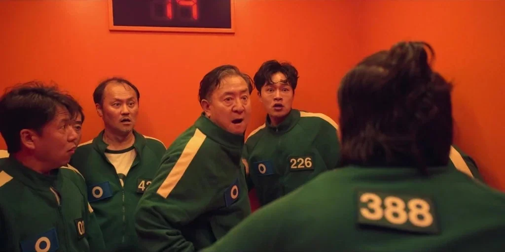 Squid Game (2024) movie still: medium — The interior of an elevator with bright orange walls and a red digital display. Five men in green numbered…