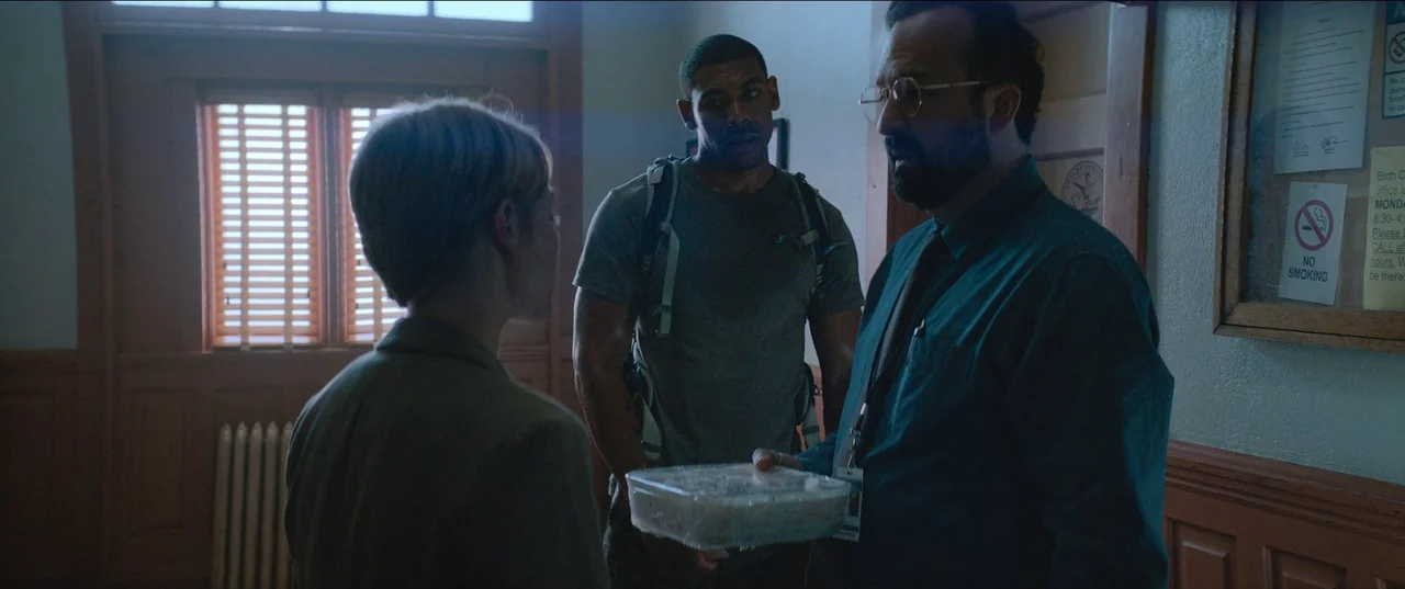 Rebel Ridge (2024) movie still: medium — An interior hallway with wooden wainscoting features a cork bulletin board on the right. Three people…