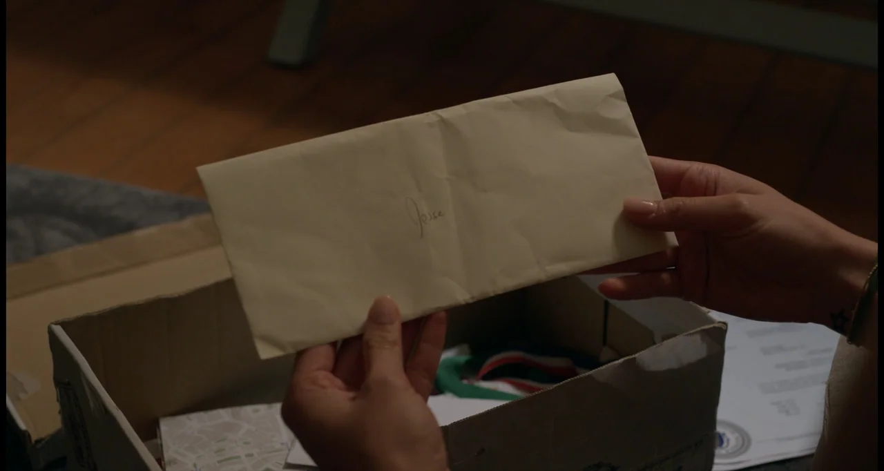 One True Loves (2023) movie still: closeup — A cardboard box sits on a wooden floor inside a room. Hands hold an off-white paper envelope with the…