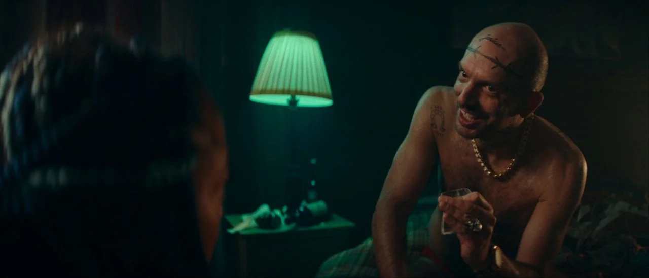 Archenemy (2020) movie still: medium — A dimly lit room features a bedside table with bottles and a green-shaded lamp. The shirtless man with facial…