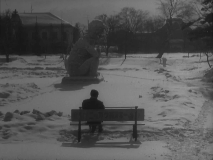 The Idiot (1951) movie still: wide — A snow-covered public park features a stone creature statue and sparse bare trees. A man sits alone on a wooden…