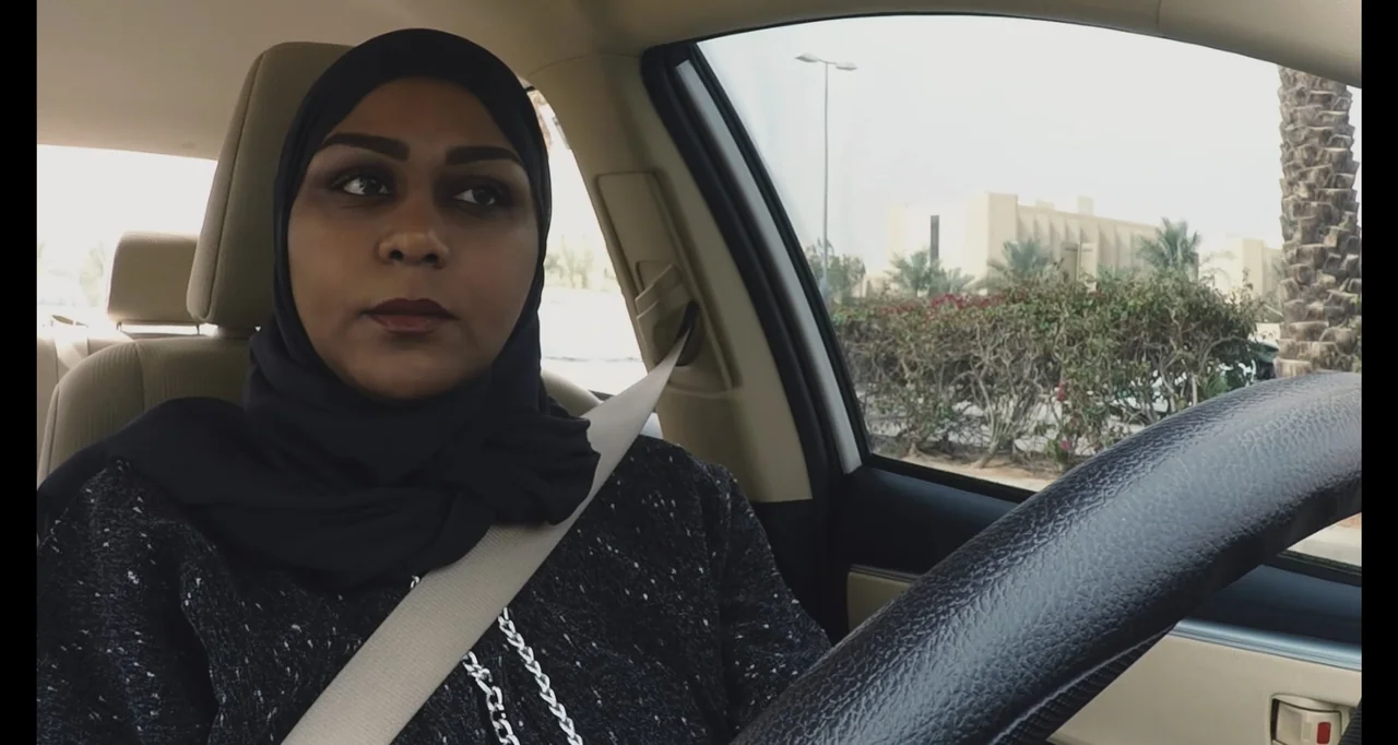 Saudi Women's Driving School (2019) movie still: medium — The interior of a moving car shows a woman behind the wheel. A view of palm trees and a…