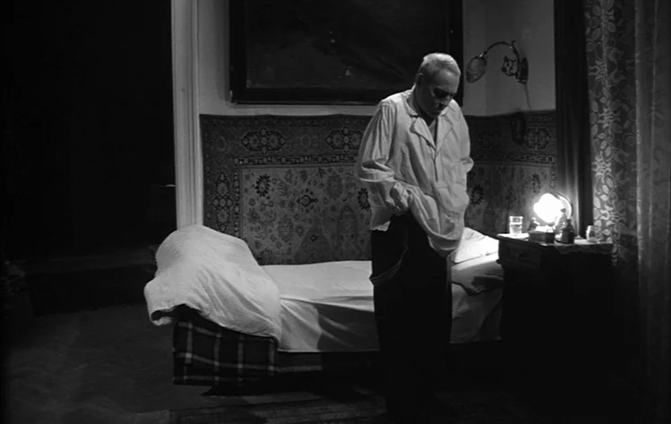 Werckmeister Harmonies (2000) movie still: medium — A dimly lit room features a single bed against a wall decorated with tapestry. An elderly man…