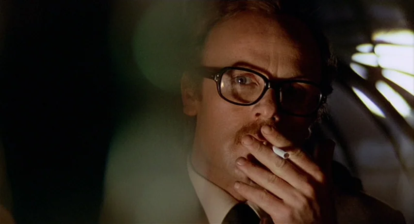 The Fifth Cord (1971) movie still: closeup — A man in a tie occupies a dimly lit space with dark surroundings. The subject holds a lit cigarette to…