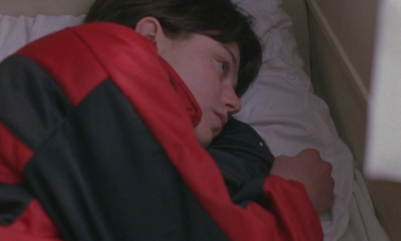 Rosetta (1999) movie still: closeup — A close-up view shows a person lying in a bed with white linens. The subject rests their head on a pillow,…