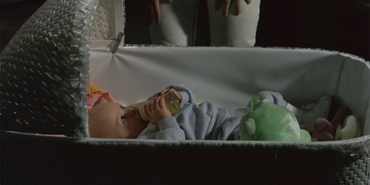 El sótano (2024) movie still: medium — A wicker infant crib rests in a dark room with a white interior lining. A baby in a grey onesie lies inside…