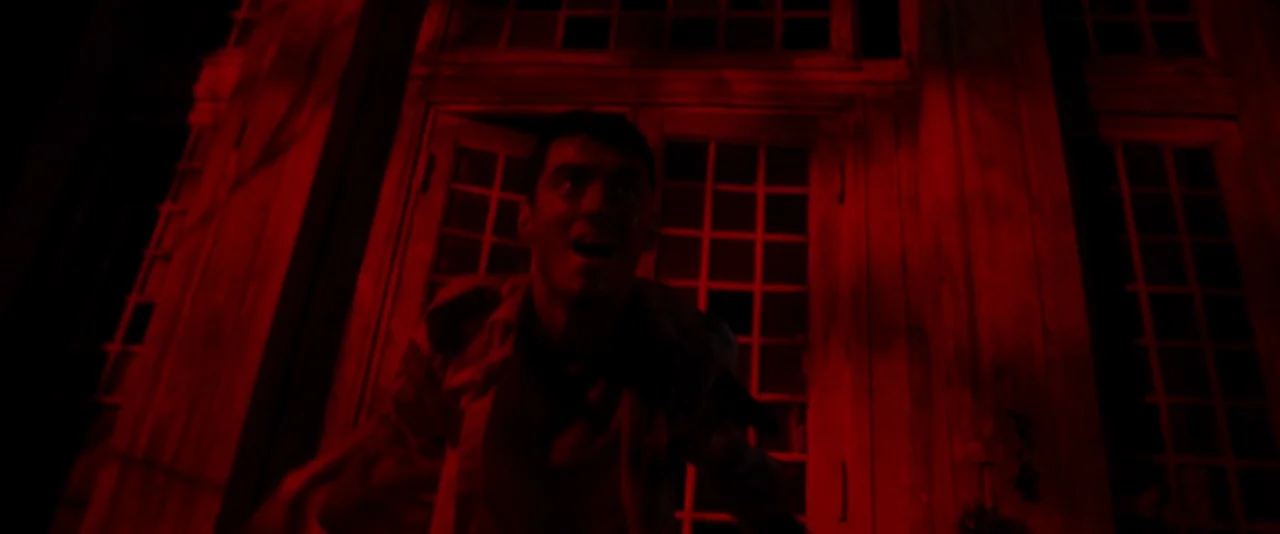Baskin (2015) movie still: medium — A man stands before a large, grid-paned window at night. Intense red light washes over the man and the building…